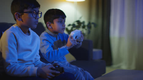 Two-Young-Boys-Sitting-On-Sofa-At-Home-Playing-With-Computer-Games-Console-On-TV-Holding-Controllers-Late-At-Night-4